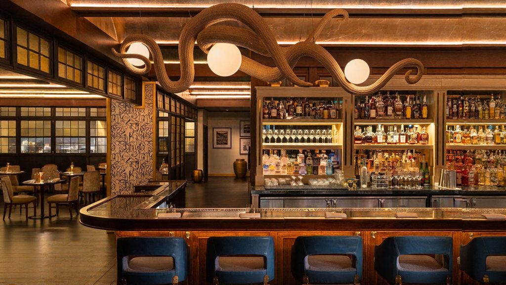 A bar with blue barstools, a well-stocked liquor shelf, and a large octopus sculpture overhead, with dining tables and chairs in the background.