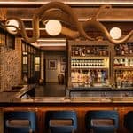 A bar with blue barstools, a well-stocked liquor shelf, and a large octopus sculpture overhead, with dining tables and chairs in the background.