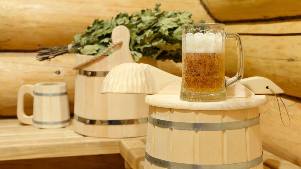 A glass of beer with foam sits on a wooden bucket in a sauna, with oak leaves and another mug in the background.