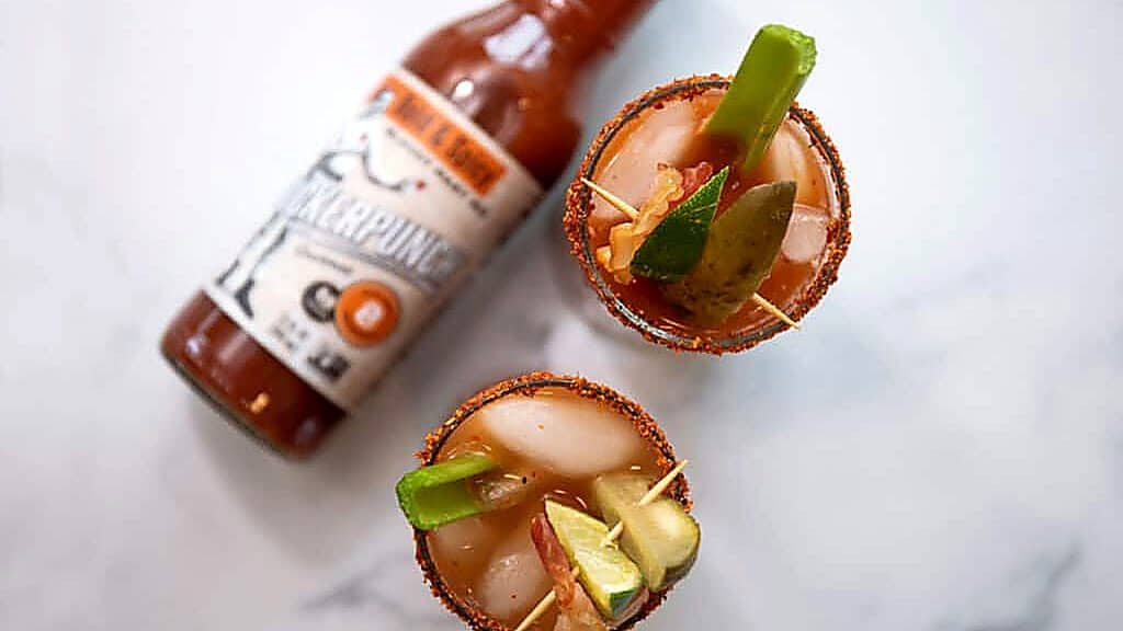 Two vegan drinks, Bloody Mary cocktails with garnishes and ice, sit next to a bottle of pepper sauce on a white marble surface.