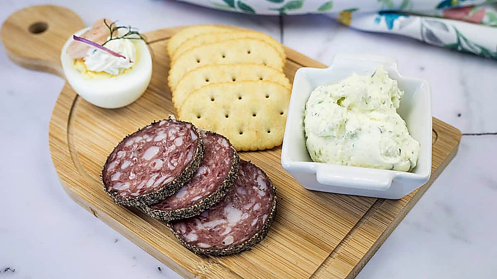 A wooden board with sliced salami, crackers, a deviled egg, and a square dish of homemade Boursin cheese.