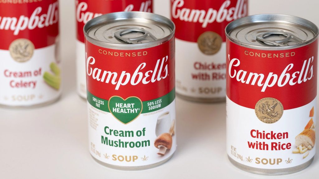 Five cans of Campbell&rsquo;s condensed canned soup, including Cream of Mushroom, Cream of Celery, and Chicken with Rice, are arranged on a white surface.