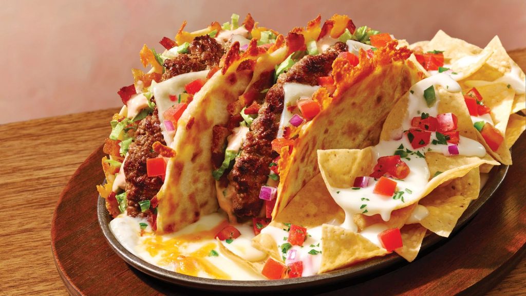 Plate with two cheesy tacos filled with seasoned meat, diced tomatoes, and onions, served with nachos topped with melted cheese and chopped vegetables.