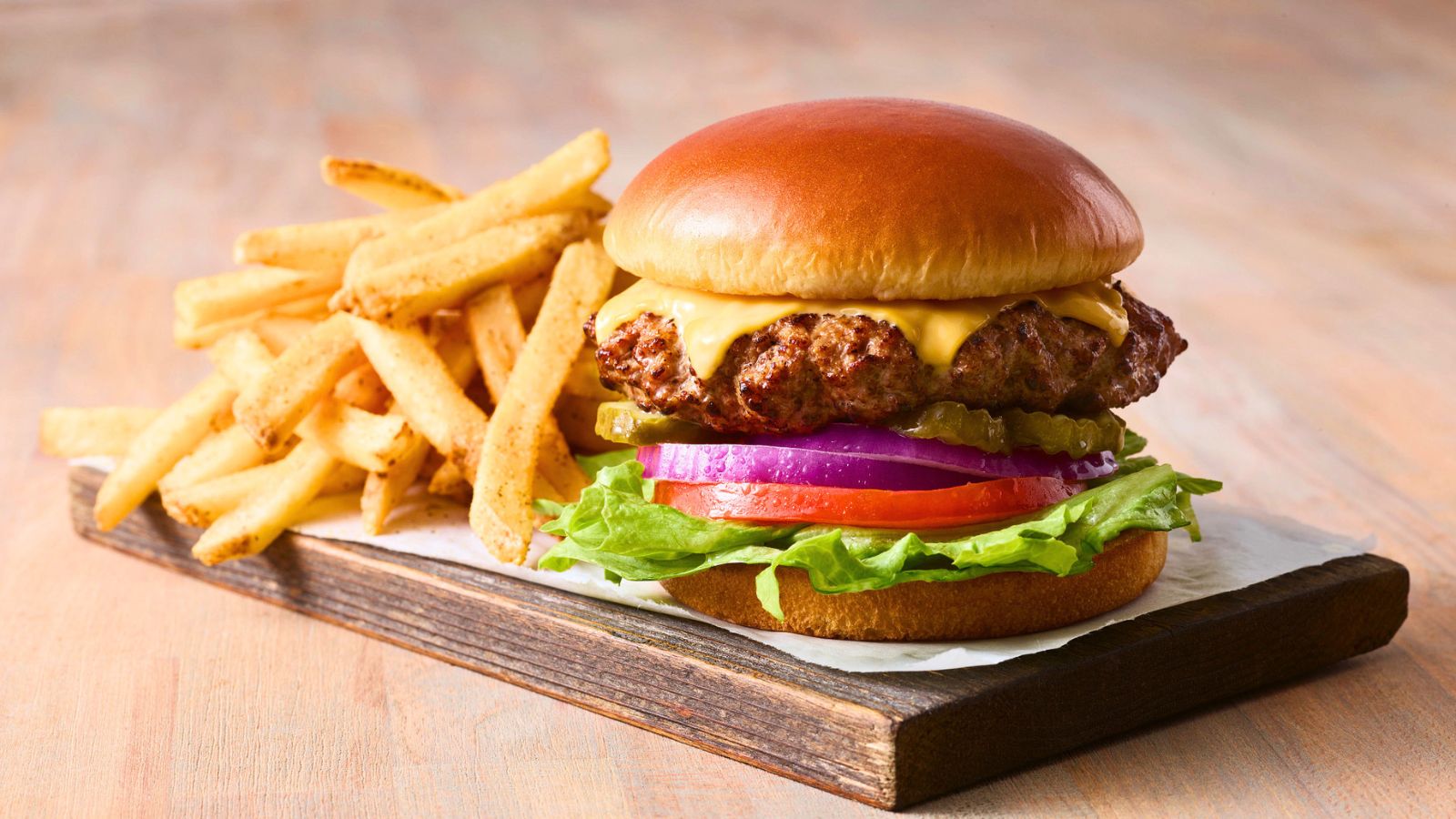 A cheeseburger with lettuce, tomato, onion, pickles, and cheese on a bun, served with a side of French fries on a wooden board.