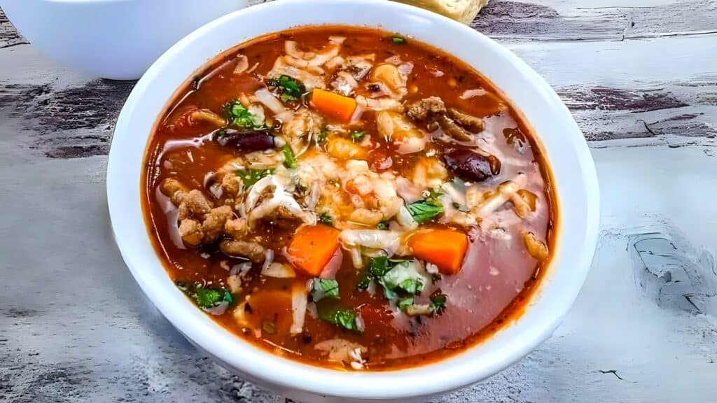 A bowl of chili with kidney beans, ground meat, carrots, shredded cheese, and chopped herbs, served on a rustic wooden surface.