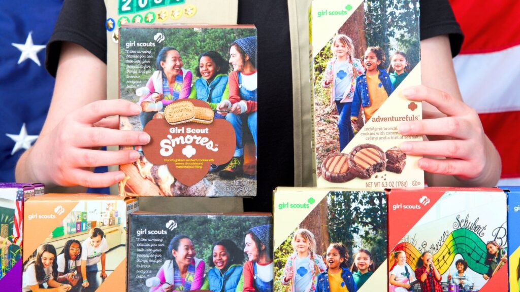 Person holding boxes of Girl Scout Cookies, including varieties like S&rsquo;mores and Adventurefuls, with others displayed in front; American flag background.