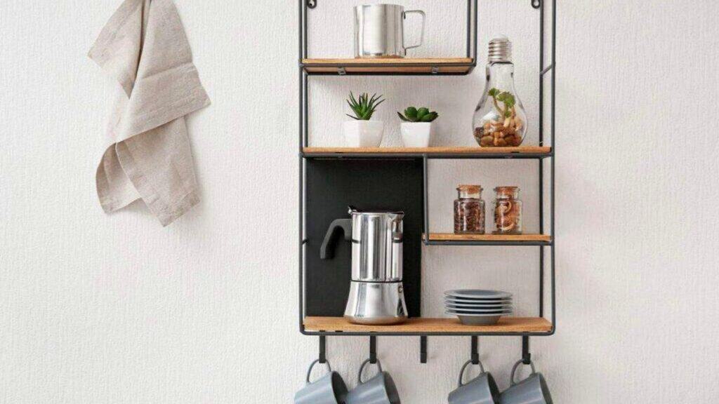Wall-mounted shelf with mugs, plates, jars, small plants, a coffee pot, a towel, and a decorative lightbulb-shaped jar&mdash;all neatly arranged against a white wall&mdash;offering inspiration for stylish coffee bar ideas.