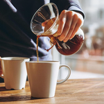 Person pouring coffee from a glass carafe into a white mug on a wooden table, with another white mug nearby, carefully preparing each coffee order.