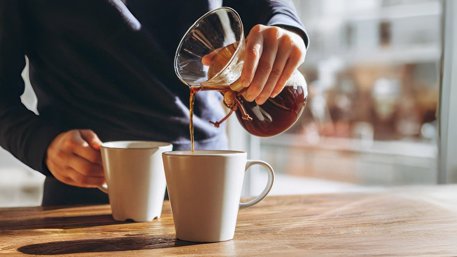 Person pouring coffee from a glass carafe into a white mug on a wooden table, with another white mug nearby, carefully preparing each coffee order.