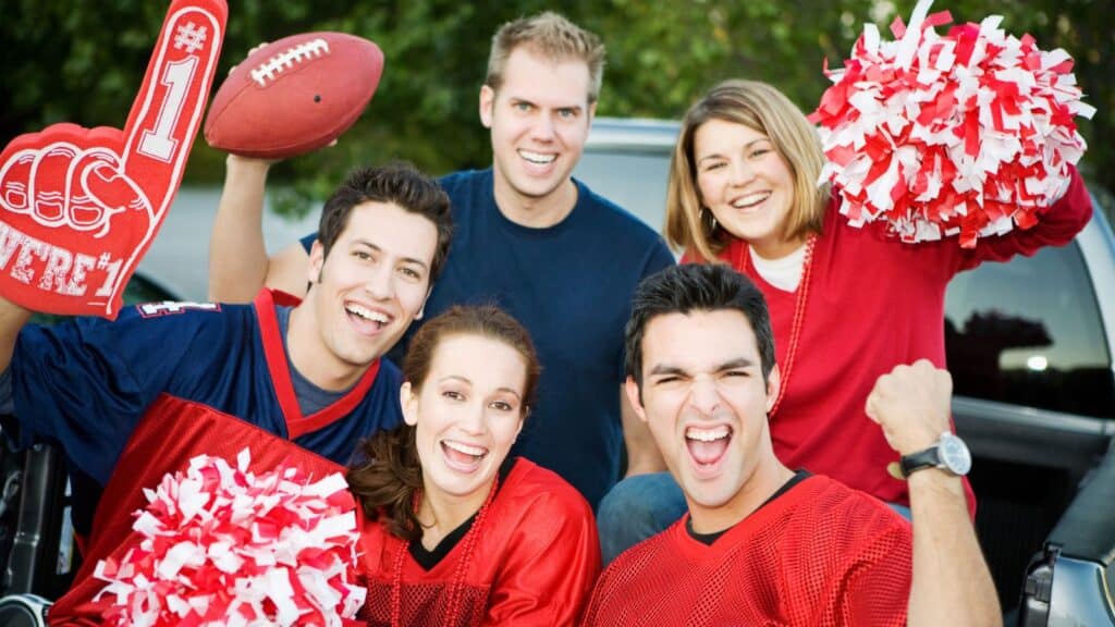 Five people wearing red and blue sports gear smile and cheer, holding pom-poms, a foam finger, and a football near a parked vehicle outdoors.