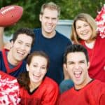 Five people wearing red and blue sports gear smile and cheer, holding pom-poms, a foam finger, and a football near a parked vehicle outdoors.