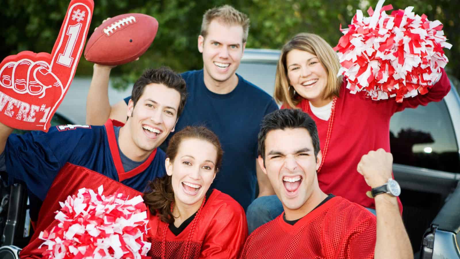 Five people wearing red and blue sports gear smile and cheer, holding pom-poms, a foam finger, and a football near a parked vehicle outdoors.