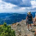 Three hikers walk along a rocky mountain trail with scenic views of valleys, hills, and cloudy sky in the background.