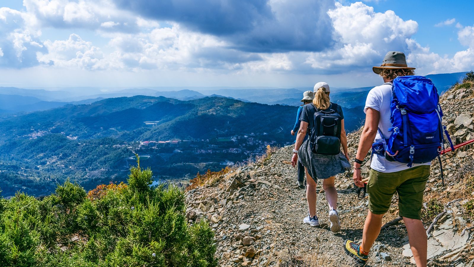 Three hikers walk along a rocky mountain trail with scenic views of valleys, hills, and cloudy sky in the background.