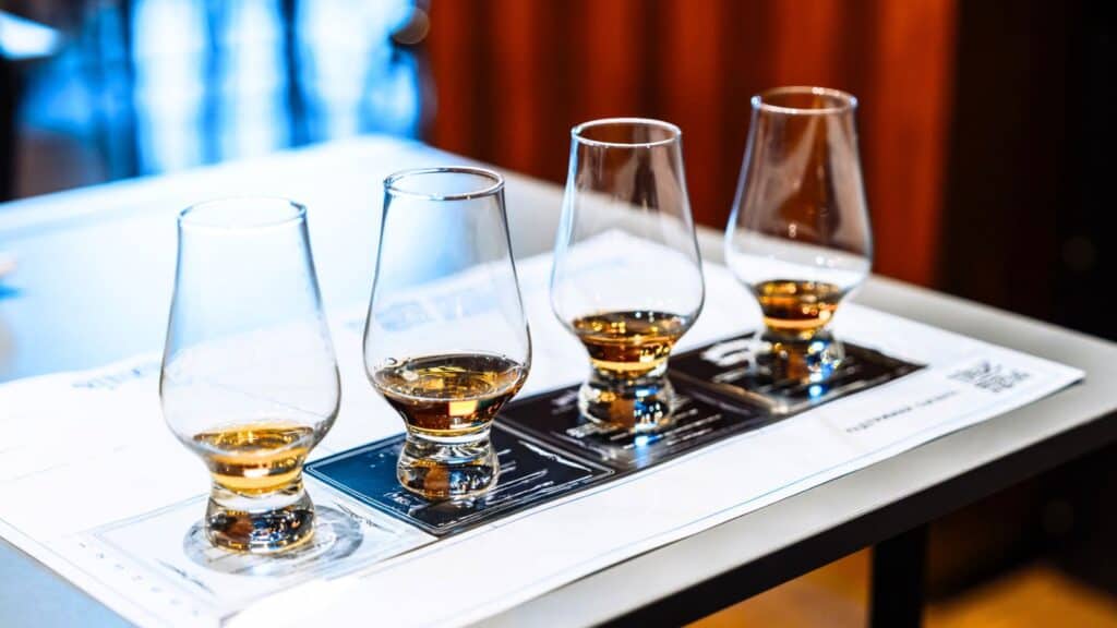 Four whiskey glasses with different levels of amber liquid are arranged in a row on a tasting mat atop a table.