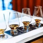 Four whiskey glasses with different levels of amber liquid are arranged in a row on a tasting mat atop a table.