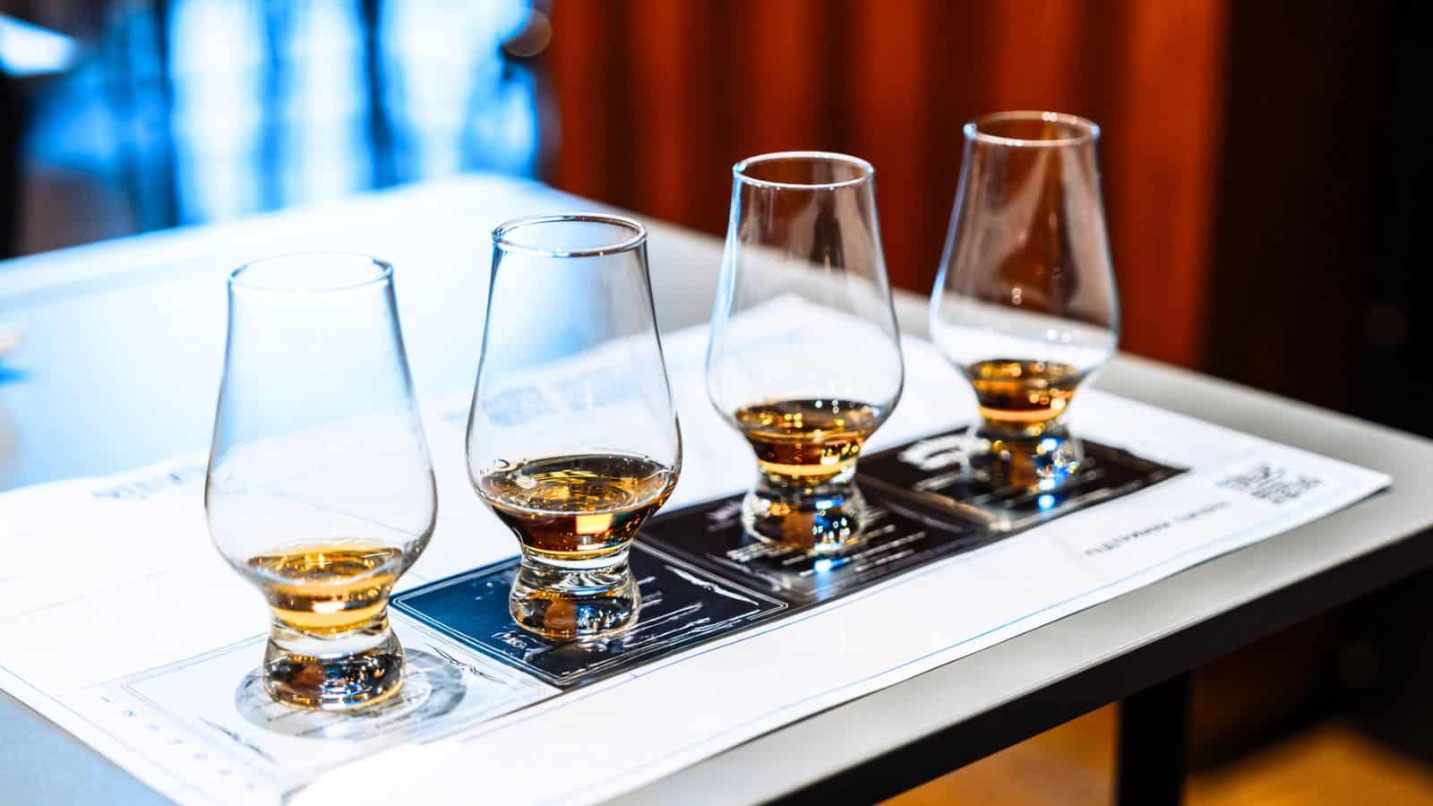 Four whiskey glasses with different levels of amber liquid are arranged in a row on a tasting mat atop a table.