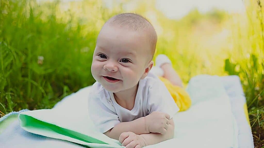 A baby in a white shirt and yellow shorts lies on a blanket on the grass, smiling at the camera on a sunny day.