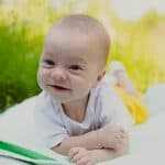 A baby in a white shirt and yellow shorts lies on a blanket on the grass, smiling at the camera on a sunny day.