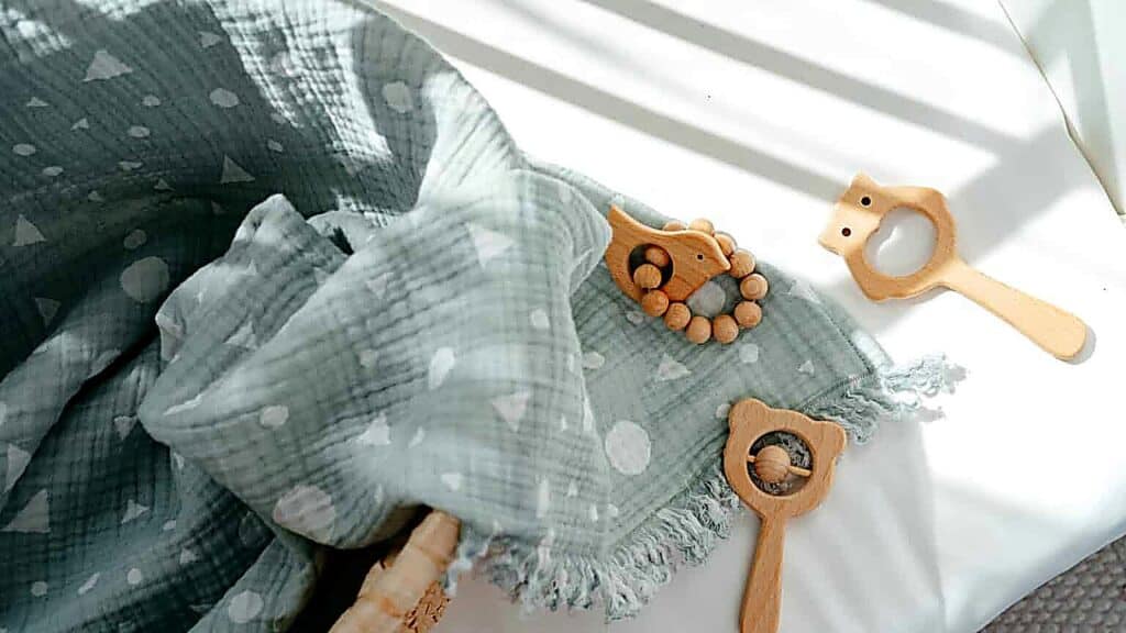 Soft green baby blanket with geometric patterns lies next to three wooden baby toys on a white surface in natural light.