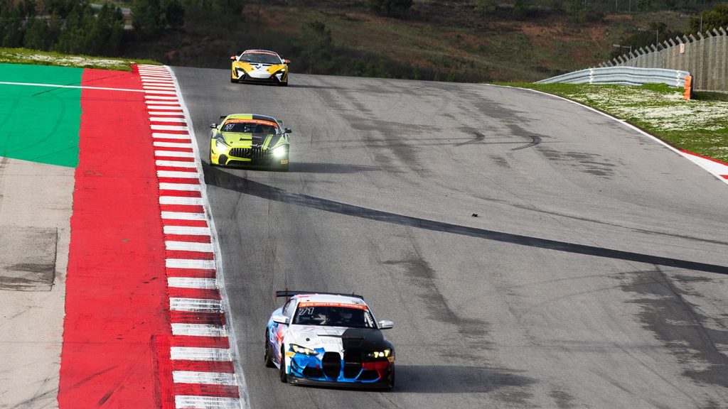 Three Formula 1 race cars drive on a curving racetrack with red and white curbs. The leading car is followed by two others, with tire marks visible on the track surface.