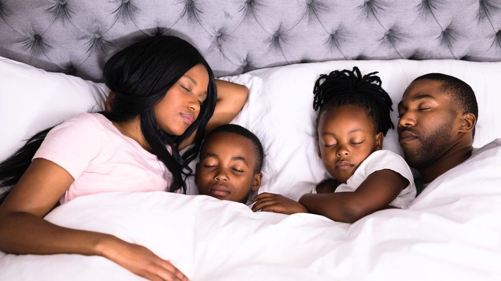 A family of four sleeps together in a bed, with two adults and two children lying under white blankets and resting peacefully, embracing healthy habits for a good night’s sleep.