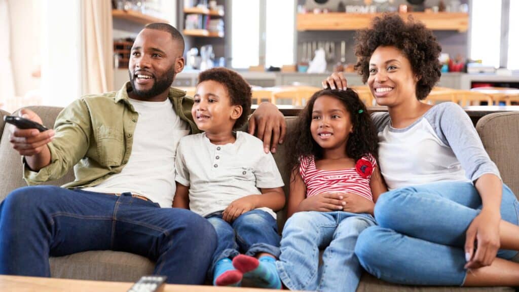 A family of four sits on a couch, smiling and watching TV together in a modern living room.