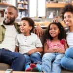 A family of four sits on a couch, smiling and watching TV together in a modern living room.