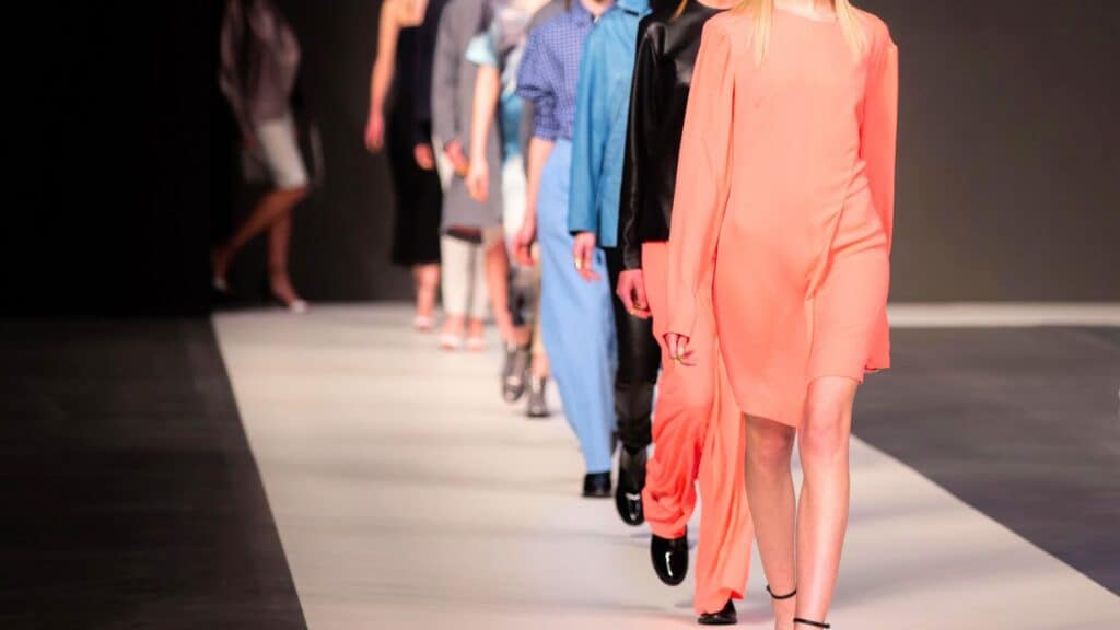 Models walk down a runway wearing various outfits during a fashion show, with a focus on the leading model in a light coral dress.