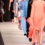 Models walk down a runway wearing various outfits during a fashion show, with a focus on the leading model in a light coral dress.