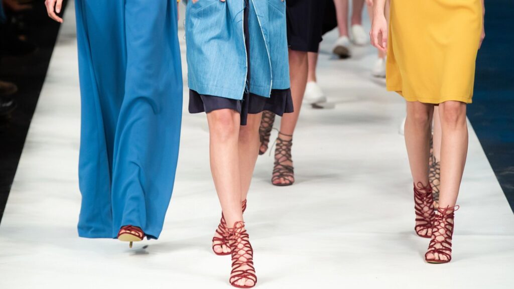 Models walk down a runway wearing brightly colored clothing and red strappy heels during a fashion show.