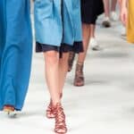 Models walk down a runway wearing brightly colored clothing and red strappy heels during a fashion show.
