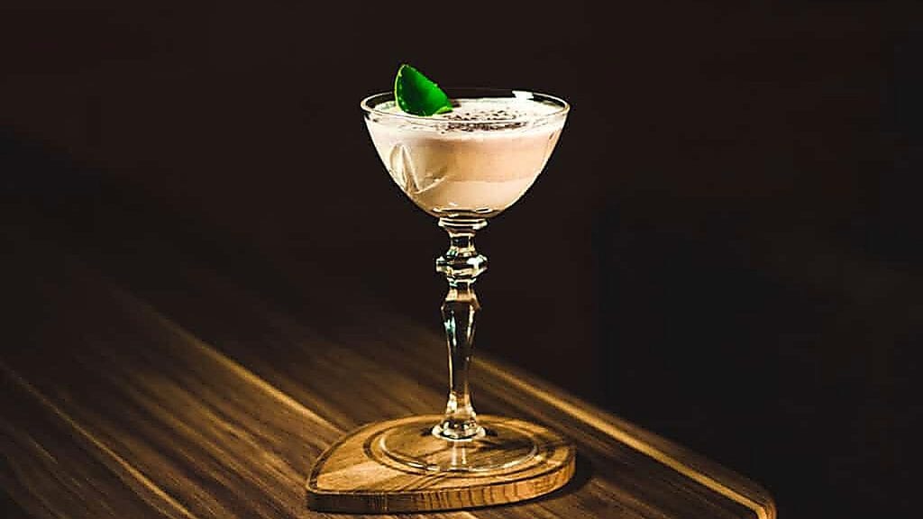 A cocktail with a frothy top and a green garnish is served in a stemmed glass on a wooden coaster, placed on a wooden surface—a sophisticated addition to any selection of vegan drinks.