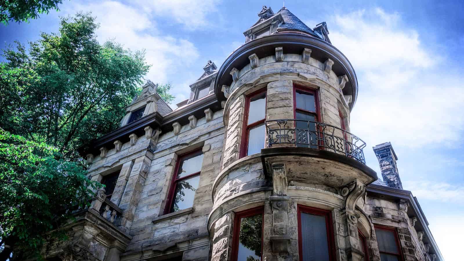 Stone mansion with arched windows, a rounded turret, balcony, and pointed roof, viewed from below against a blue sky with green tree branches on the left.