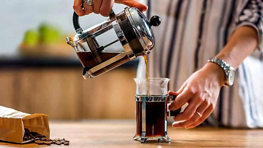 A person pours coffee from a French press into a glass mug on a wooden table, with a bag of coffee beans nearby&mdash;capturing the essence of cozy coffee bar ideas for your home.