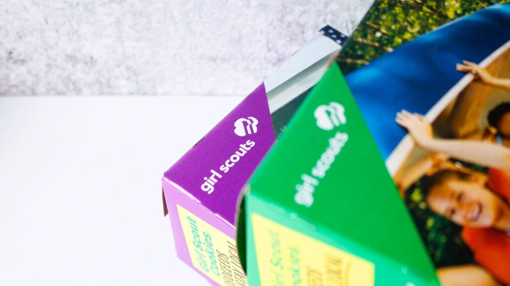 Close-up of two Girl Scouts cookie boxes, one purple and one green, partially open on a white surface with part of a smiling girl visible on the green box.