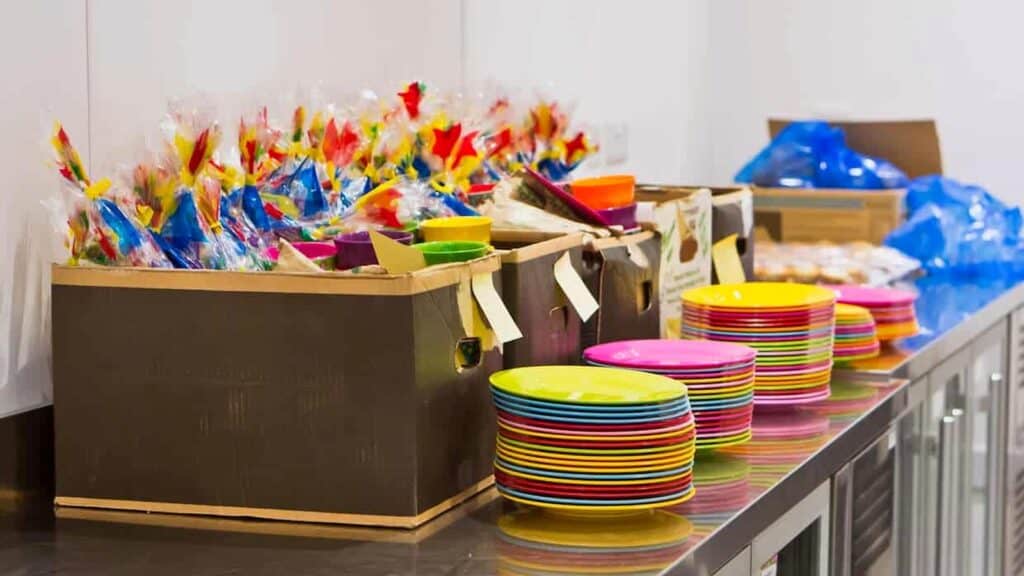 Colorful plastic plates stacked beside cardboard boxes filled with wrapped party favors on a stainless steel countertop.