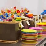 Colorful plastic plates stacked beside cardboard boxes filled with wrapped party favors on a stainless steel countertop.