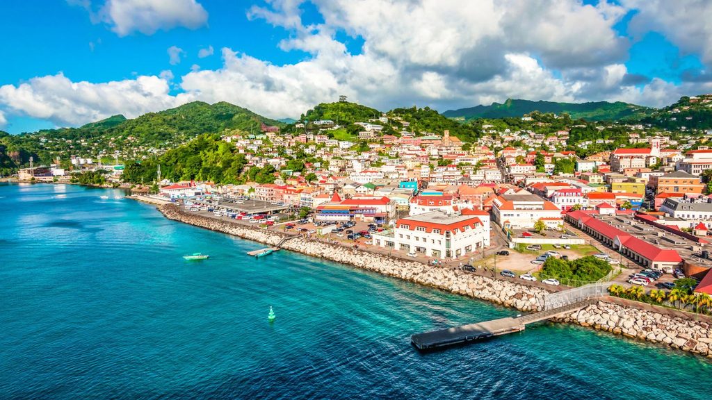 A coastal town with colorful buildings, green hills in the background, boats in blue water, and a partly cloudy sky above.