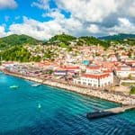 A coastal town with colorful buildings, green hills in the background, boats in blue water, and a partly cloudy sky above.