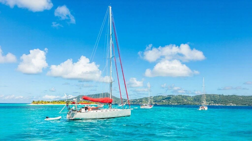 A sailboat with a red cover floats on clear turquoise water near an island, with hills and two other sailboats visible in the background under a blue sky with scattered clouds.