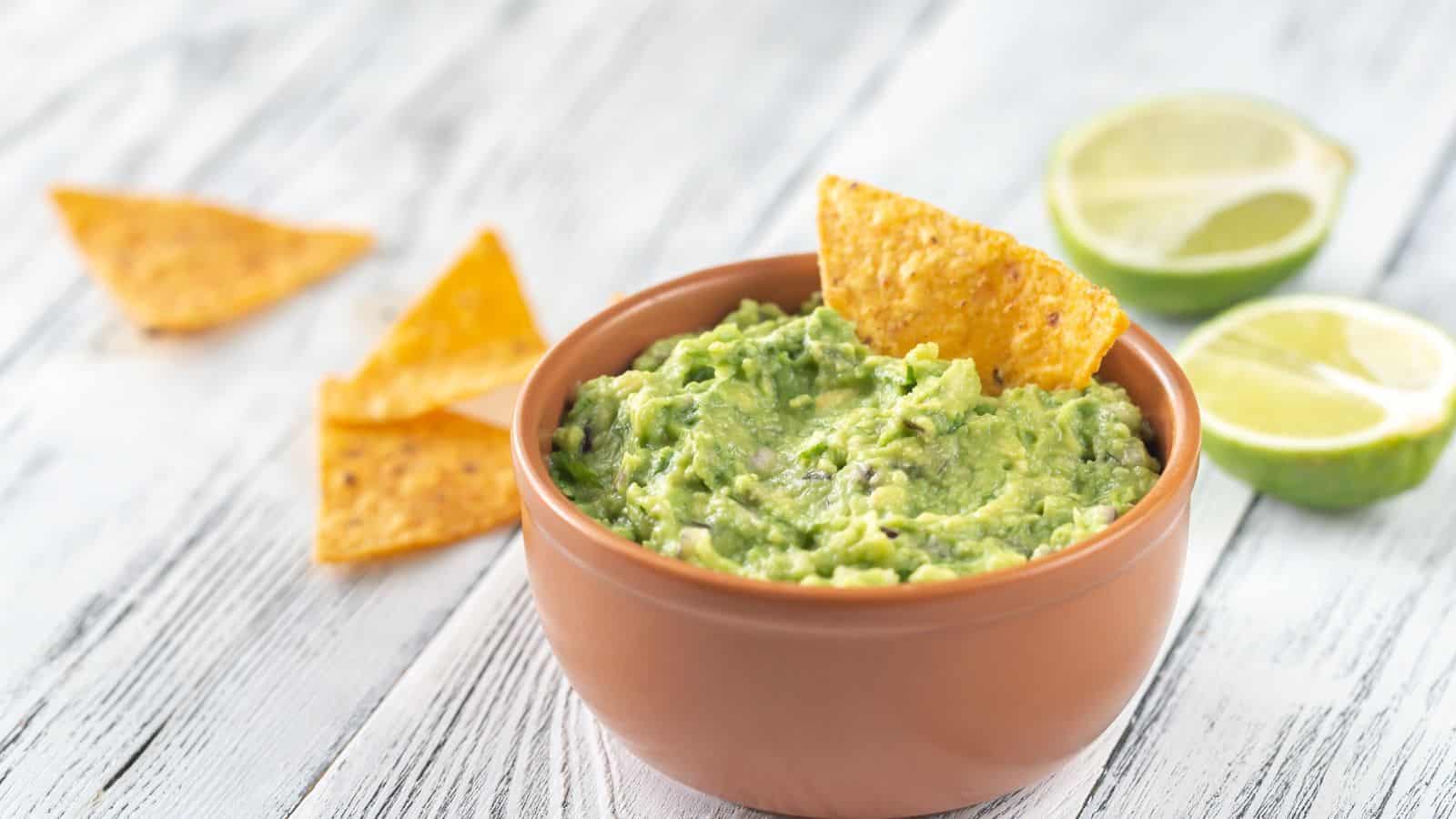 A bowl of guacamole with a tortilla chip dipped in, surrounded by tortilla chips and lime halves on a light wooden surface.