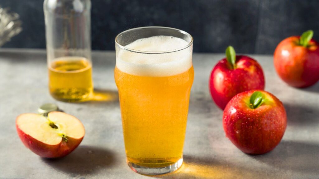 A glass of apple cider with foam sits on a table next to a bottle of cider and three red apples, one of which is sliced in half.
