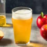 A glass of apple cider with foam on top sits on a table next to a half-full bottle and three old apples, one of which is sliced.