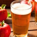 A glass of apple cider sits on a wooden surface next to fresh red apples, with another glass of cider in the background.