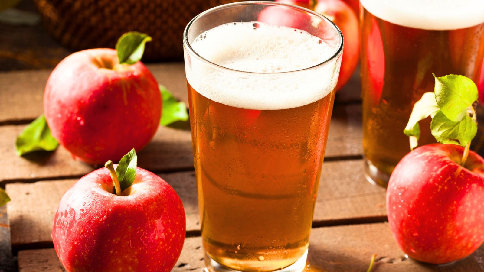 A glass of apple cider sits on a wooden surface next to fresh red apples, with another glass of cider in the background.
