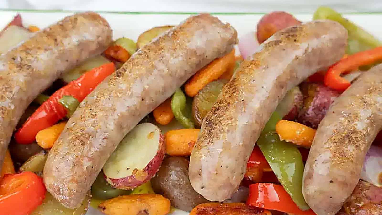 Four cooked sausages served on a bed of roasted vegetables, including carrots, red potatoes, green bell peppers, and red bell peppers.