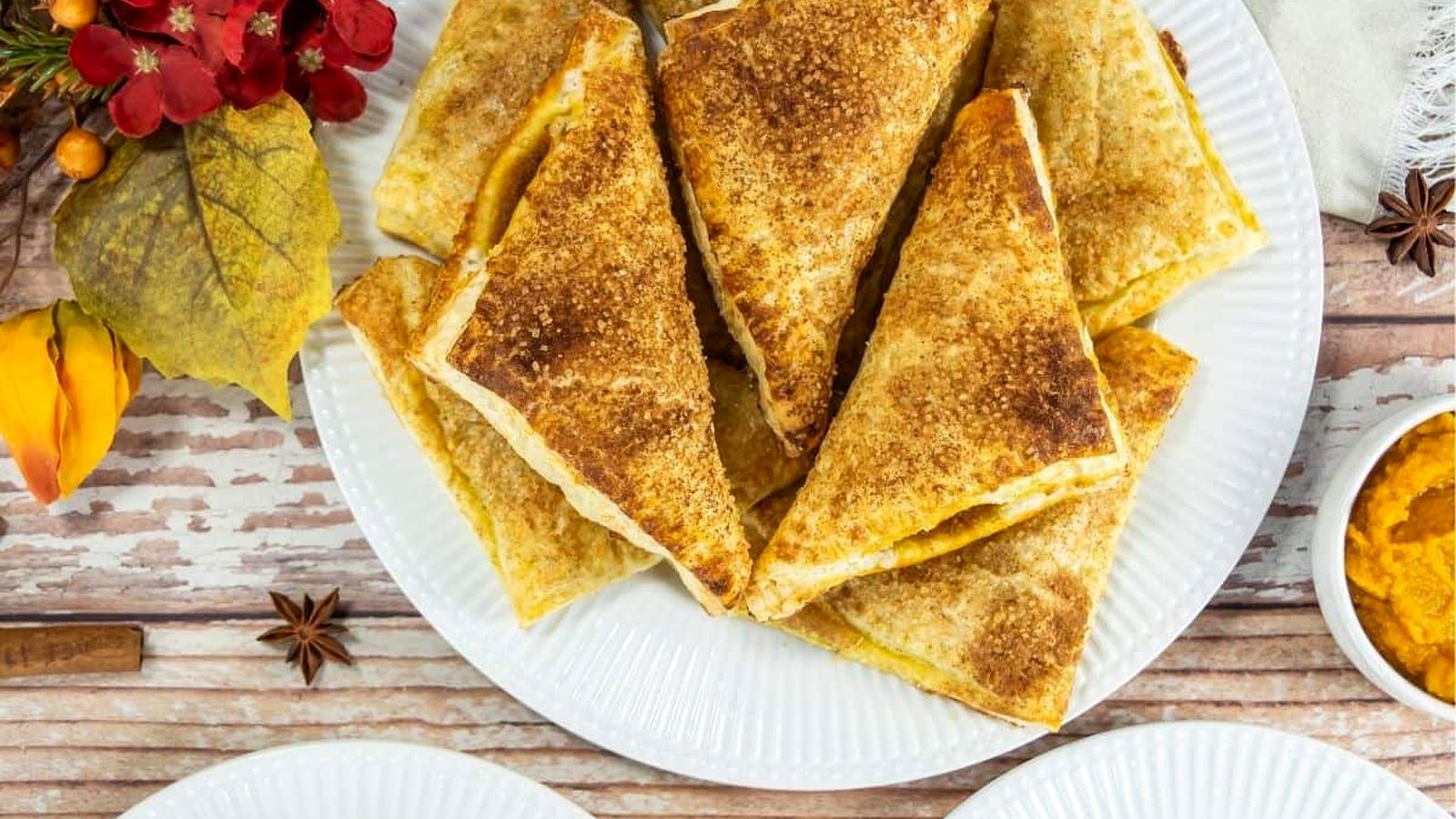 A plate of Pumpkin Turnovers arranged in a stack, with autumn-themed decorations and a small bowl of orange spread nearby.