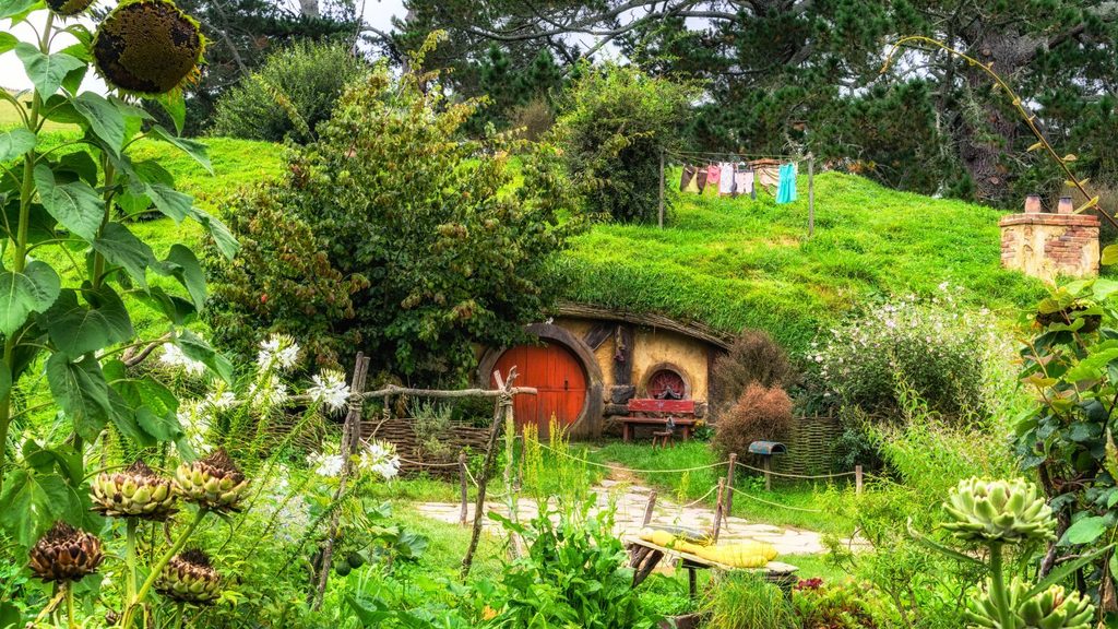 A small, round-door house built into a grassy hillside evokes the charm of Hobbit Day, surrounded by lush greenery, colorful flowers, and hanging laundry gently swaying on a clothesline.