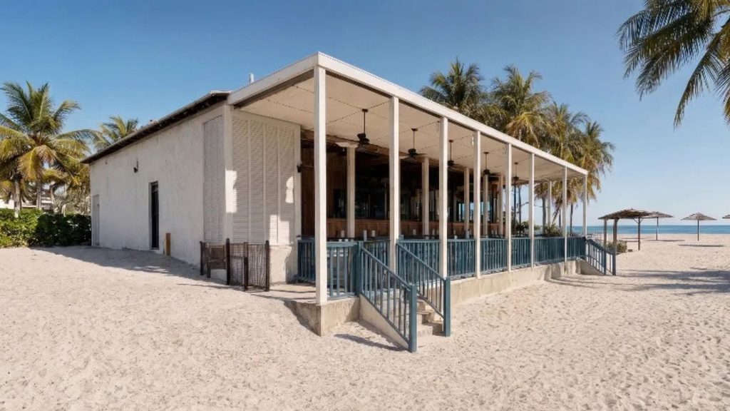 A white beachside building with a covered porch and blue railings sits on sandy ground, surrounded by palm trees under a clear sky.